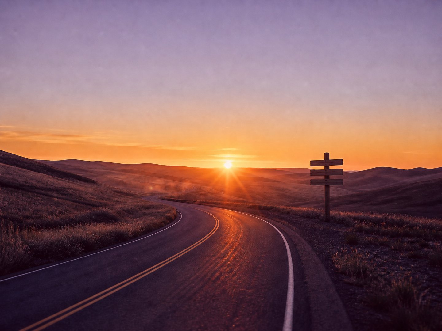 An empty two-lane road curving toward a sunrise over open hills, with a three-arm wooden signpost standing beside it